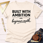 Women's funny woodworking graphic tee shirt for female woodworkers, makers, and DIY builders — Sawdust & Ponytails. Cream t-shirt with "Built With Ambition And Beginner Math" in black text.