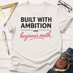 Women's funny woodworking graphic tee shirt for female woodworkers, makers, and DIY builders — Sawdust & Ponytails. White cotton tee with "Built With Ambition And beginner math" in black and pink text.