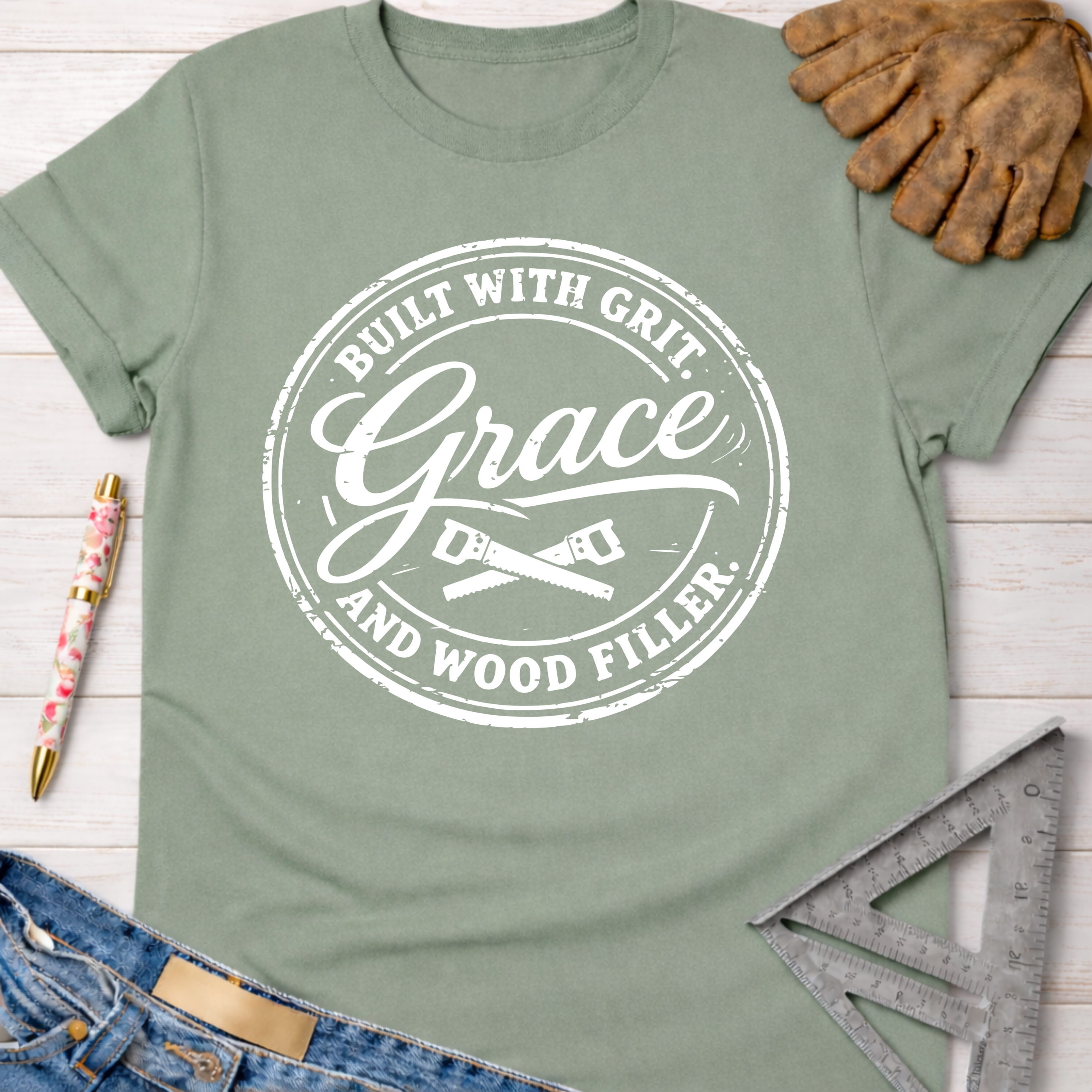 Women's funny woodworking graphic tee shirt for female woodworkers, makers, and DIY builders — Sawdust & Ponytails. Sage green cotton tee with "Built With Grit, Grace, and Wood Filler" graphic.