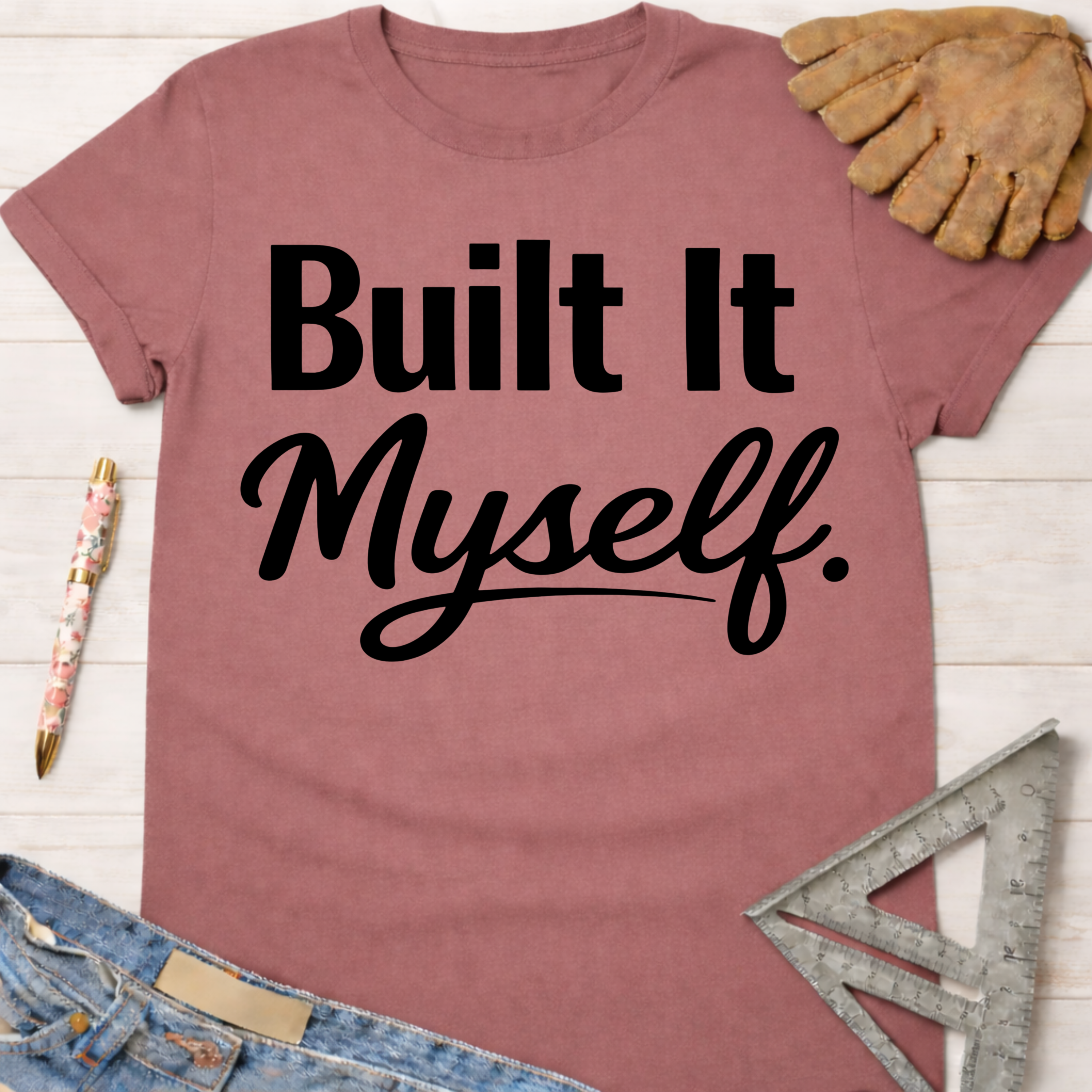 Women's funny woodworking graphic tee shirt for female woodworkers, makers, and DIY builders — Sawdust & Ponytails. Mauve pink cotton t-shirt with "Built It Myself." in black text, styled with a work glove, pen, and ruler.