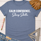Women's funny woodworking graphic tee shirt for female woodworkers, makers, and DIY builders — Sawdust & Ponytails. Blue cotton t-shirt with "Calm Confidence. Sharp Skills." in white text, next to work gloves and a ruler.