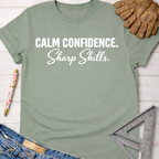 Sage green women's tee with "Calm Confidence. Sharp Skills." graphic, jeans, work glove, and ruler. Sawdust & Ponytails.