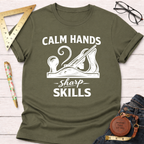 Olive green "Calm Hands Sharp Skills" t-shirt with a white hand plane graphic, folded on a white wood surface.