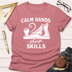 Dusty rose t-shirt with "Calm Hands Sharp Skills" text and a hand plane graphic, styled with jeans and tools.