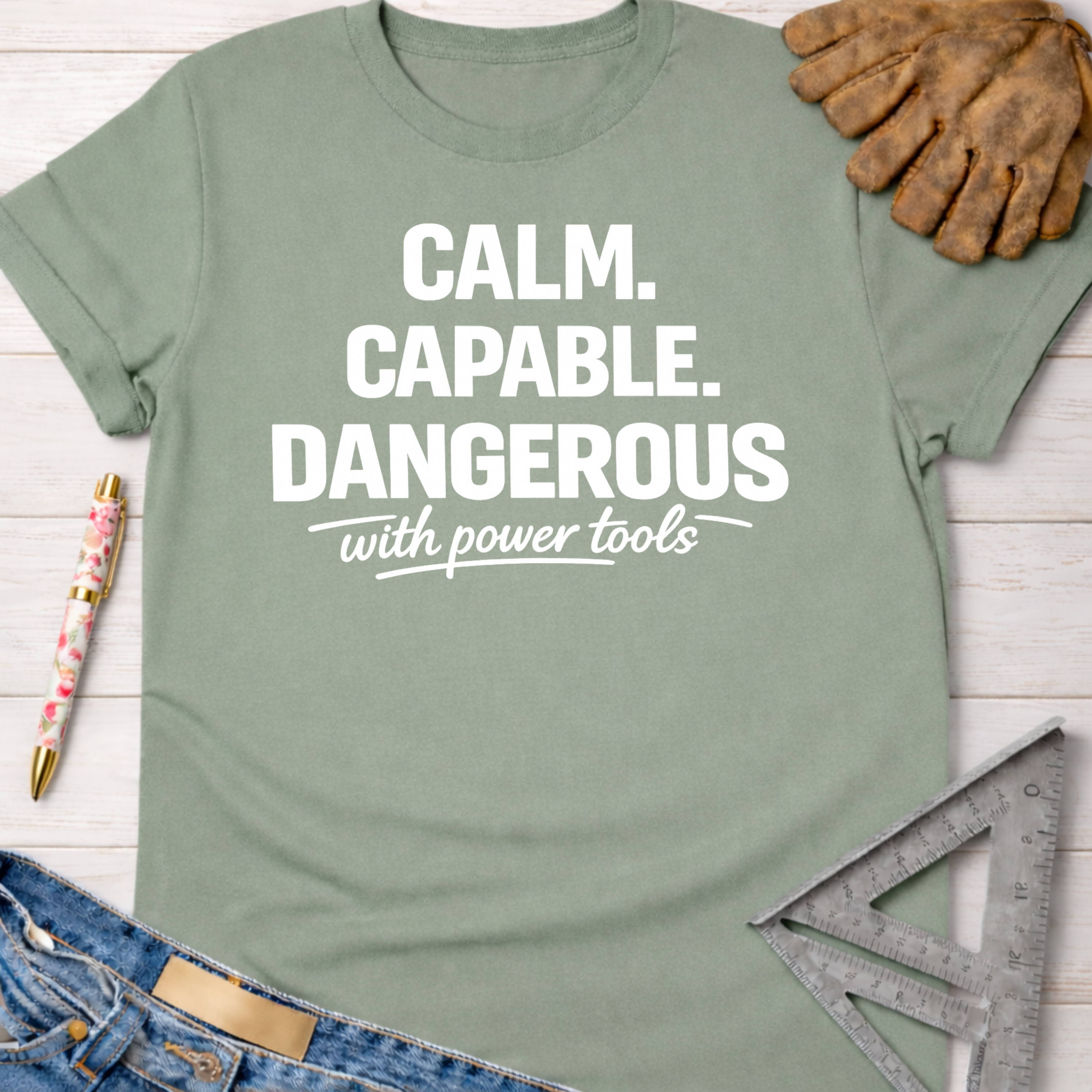 Women's funny woodworking graphic tee shirt for female woodworkers, makers, and DIY builders — Sawdust & Ponytails. Sage green cotton tee with "Calm. Capable. Dangerous with power tools" in white text.