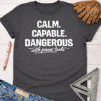 Women's funny woodworking graphic tee shirt for female woodworkers, makers, and DIY builders — Sawdust & Ponytails. Dark gray cotton tee with "CALM. CAPABLE. DANGEROUS with power tools" in white text.