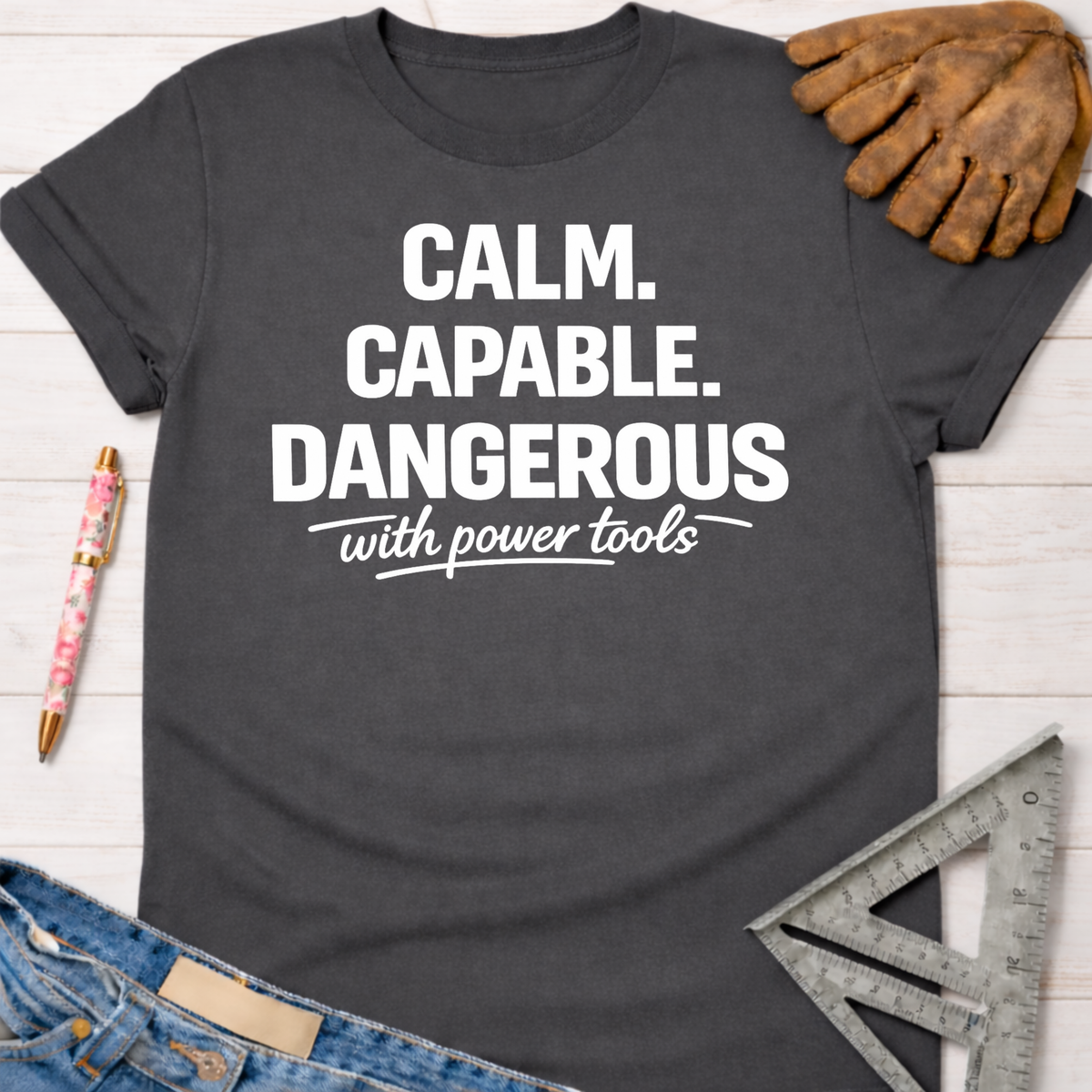 Women's funny woodworking graphic tee shirt for female woodworkers, makers, and DIY builders — Sawdust & Ponytails. Dark gray cotton tee with "CALM. CAPABLE. DANGEROUS with power tools" in white text.