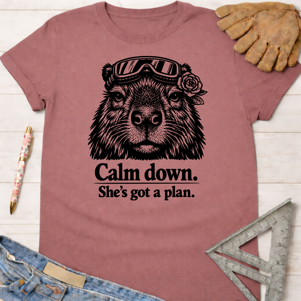 Women's funny woodworking graphic tee shirt for female woodworkers, makers, and DIY builders — Sawdust & Ponytails. Mauve t-shirt with a black graphic of a capybara wearing goggles and a rose, with the text "Calm down. She's got a plan.