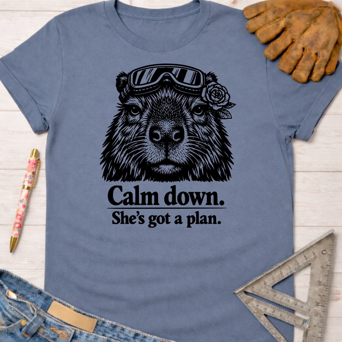 Women's funny woodworking graphic tee shirt for female woodworkers, makers, and DIY builders — Sawdust & Ponytails. Blue cotton t-shirt with a black graphic of a capybara wearing goggles and a rose, with the text "Calm down. She's got a plan.