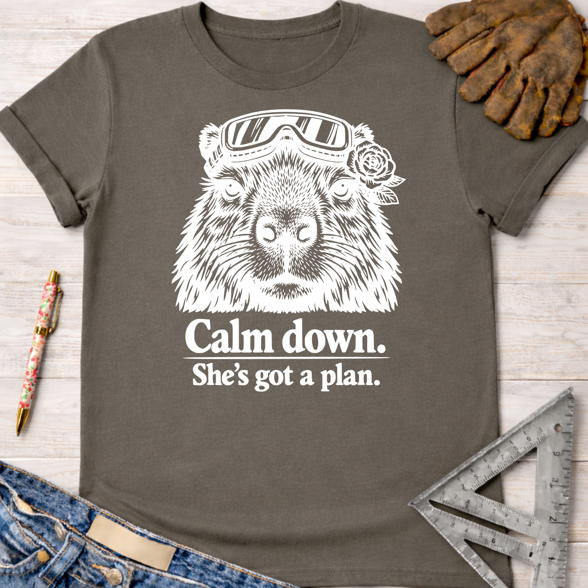 Women's funny woodworking graphic tee shirt for female woodworkers, makers, and DIY builders — Sawdust & Ponytails. Dark gray cotton tee with a white graphic of a capybara wearing goggles and a rose, with the text "Calm down. She's got a plan.