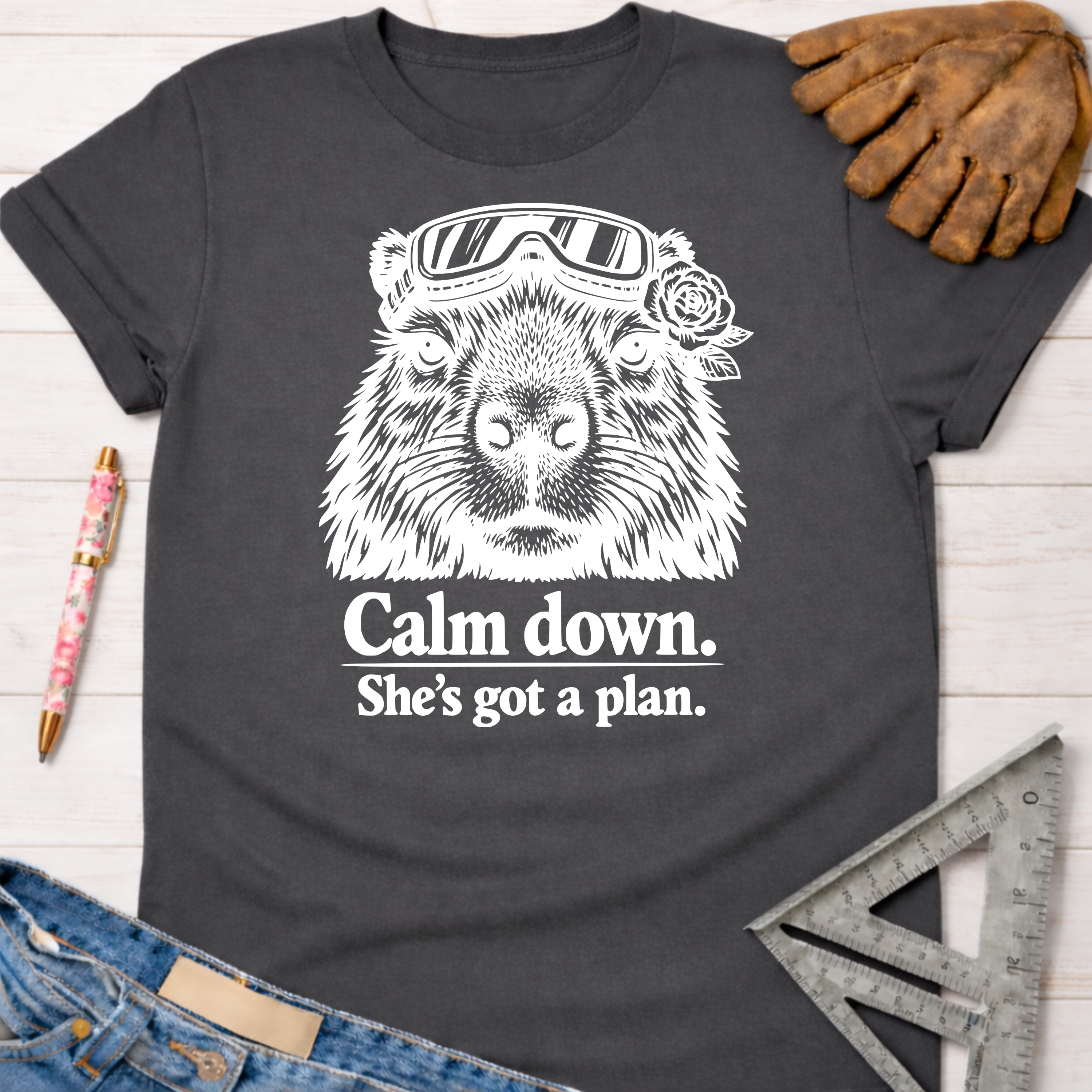 Women's funny woodworking graphic tee shirt for female woodworkers, makers, and DIY builders — Sawdust & Ponytails. Dark gray cotton tee with a white graphic of a capybara wearing goggles and a rose, with the text "Calm down. She's got a plan.