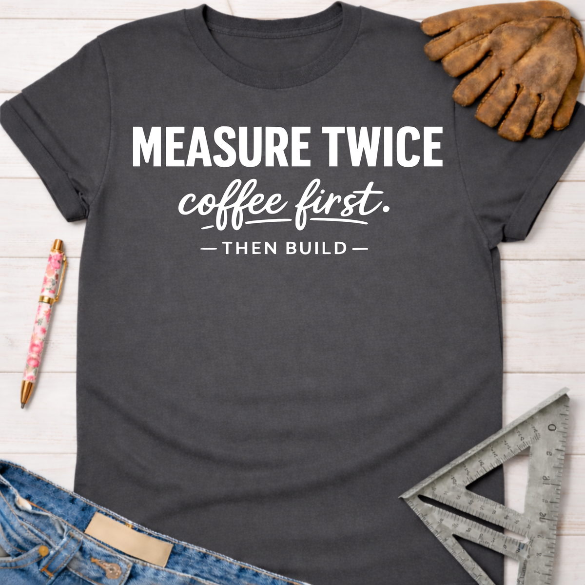 Women's funny woodworking graphic tee shirt for female woodworkers, makers, and DIY builders — Sawdust & Ponytails. Dark gray crew neck t-shirt with "MEASURE TWICE coffee first. — THEN BUILD —" in white text, styled with jeans, a pen, and a work glove.