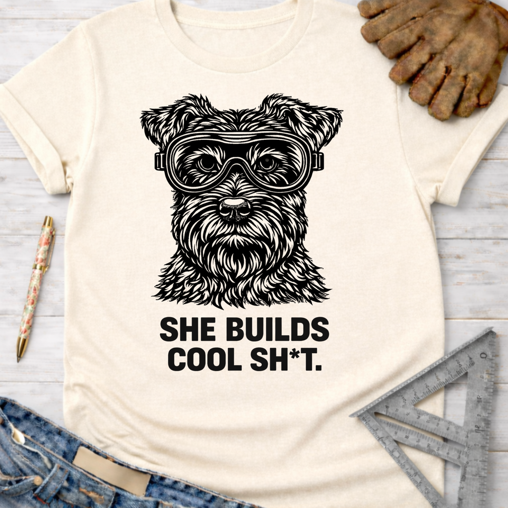 Women's funny woodworking graphic tee shirt for female woodworkers, makers, and DIY builders — Sawdust & Ponytails. Cream t-shirt with a black graphic of a schnauzer wearing safety goggles and the text "SHE BUILDS COOL SH*T.