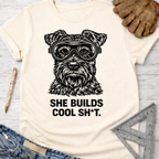 Women's funny woodworking graphic tee shirt for female woodworkers, makers, and DIY builders — Sawdust & Ponytails. Cream t-shirt with a black graphic of a schnauzer wearing safety goggles and the text "SHE BUILDS COOL SH*T.