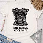 Women's funny woodworking graphic tee shirt for female woodworkers, makers, and DIY builders — Sawdust & Ponytails. White cotton tee with a black graphic of a schnauzer wearing safety goggles and the text "SHE BUILDS COOL SH*T.