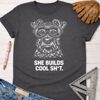 Charcoal gray women's funny woodworking graphic tee with a white dog wearing goggles and "SHE BUILDS COOL SH*T." text.