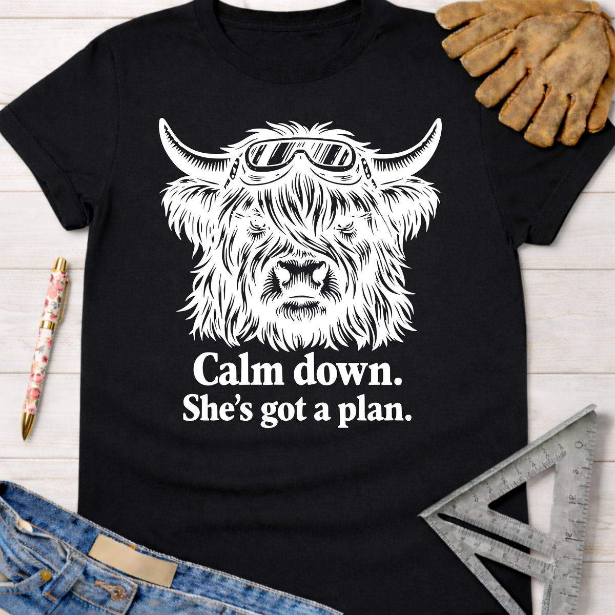 Black cotton t-shirt with a white graphic of a Highland cow wearing goggles and "Calm down. She's got a plan." Women's funny woodworking graphic tee shirt for female woodworkers, makers, and DIY builders — Sawdust & Ponytails