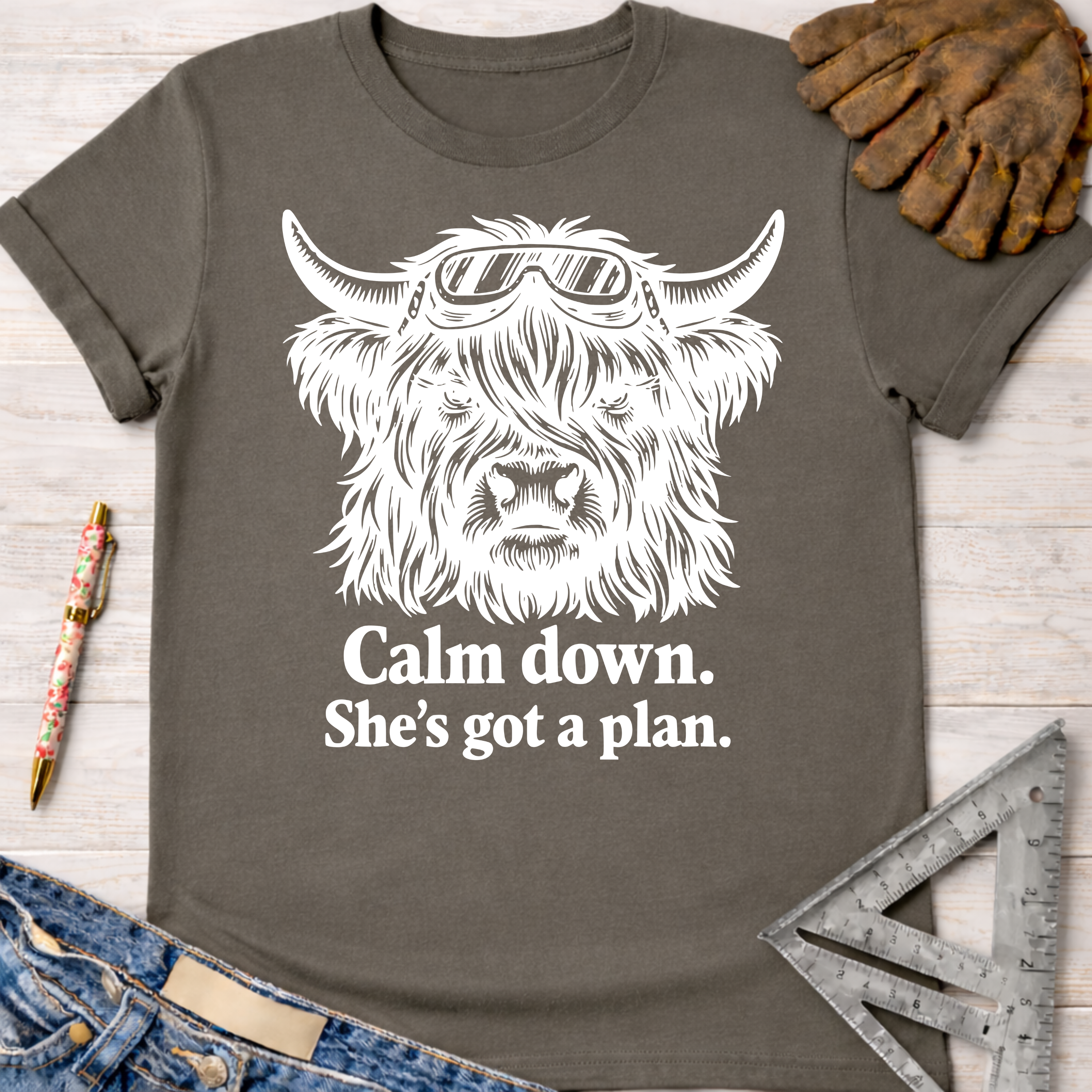 Women's funny woodworking graphic tee shirt for female woodworkers, makers, and DIY builders — Sawdust & Ponytails. Dark gray cotton tee with a white graphic of a Highland cow in safety goggles and text "Calm down. She's got a plan.