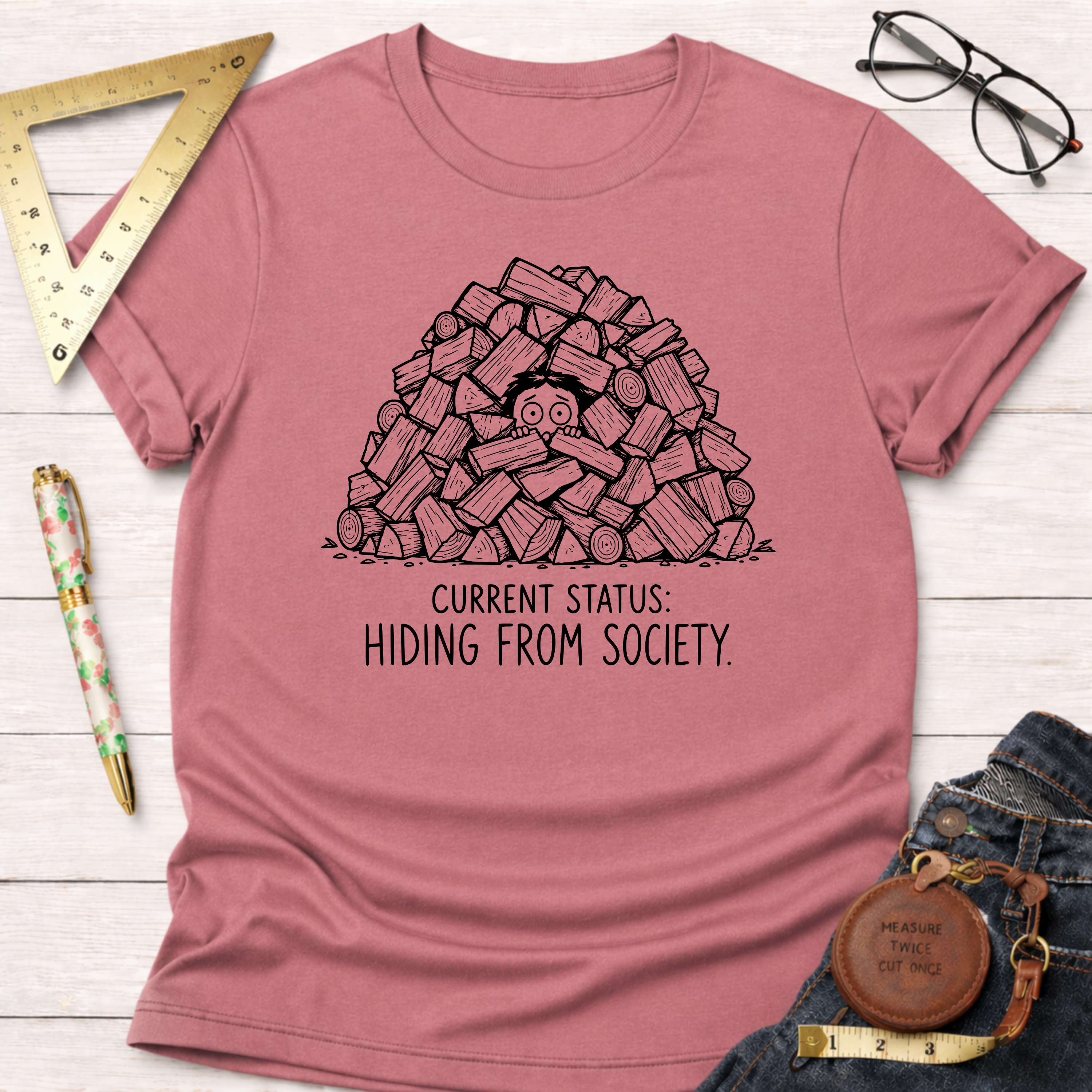 Dusty rose t-shirt with "Current Status: Hiding From Society" graphic of a person peeking from a woodpile.