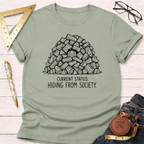 Sage green cotton t-shirt with "Current Status: Hiding From Society" text and a person peeking from a woodpile.