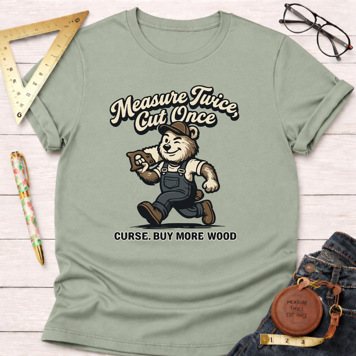 Women's funny woodworking graphic tee shirt for female woodworkers, makers, and DIY builders — Sawdust & Ponytails. Sage green cotton tee with a cartoon bear carrying wood and the text "Measure Twice, Cut Once. Curse. Buy More Wood.