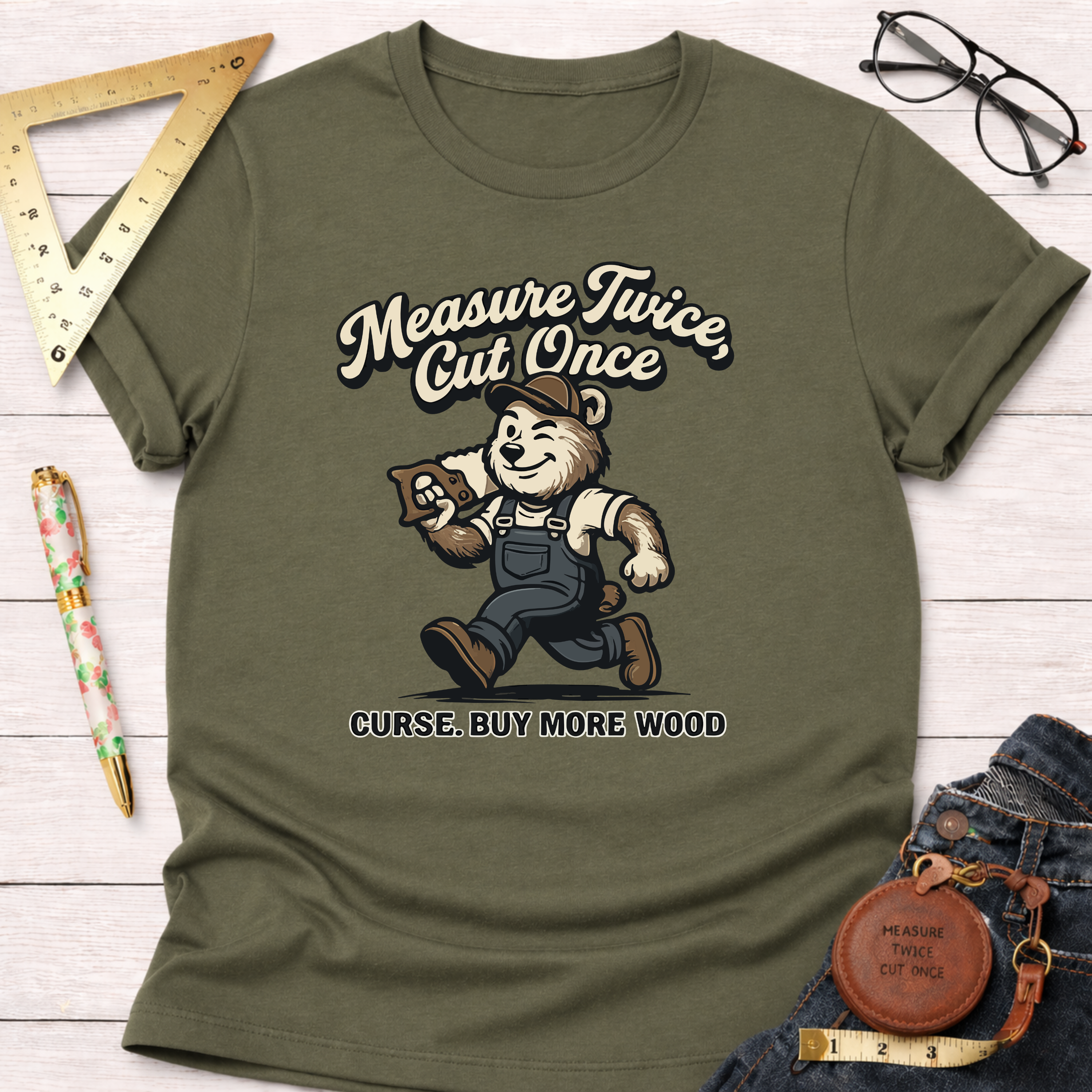 Women's funny woodworking graphic tee shirt for female woodworkers, makers, and DIY builders — Sawdust & Ponytails. Olive green cotton tee with a cartoon bear carpenter graphic and "Measure Twice, Cut Once. Curse. Buy More Wood" text.