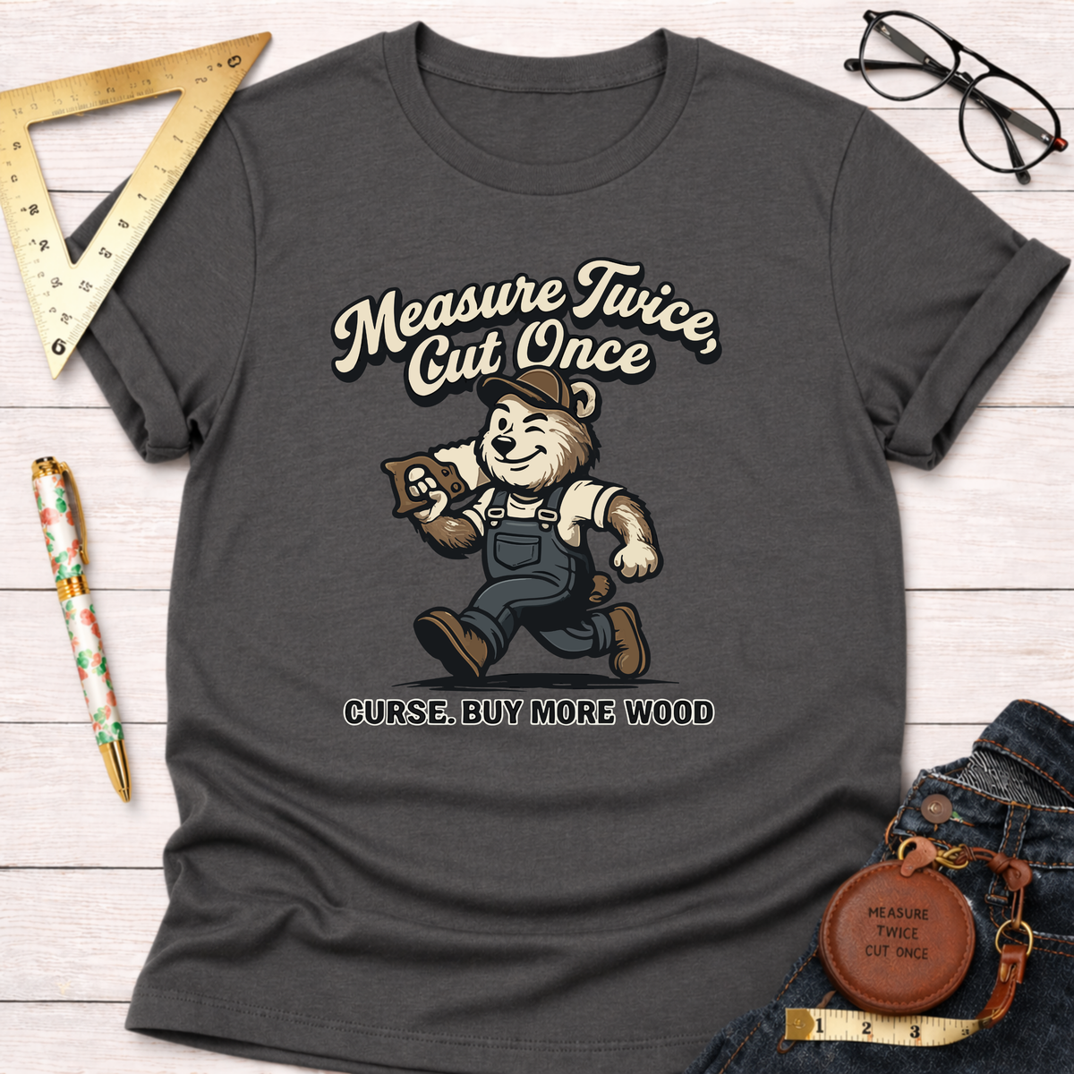 Women's funny woodworking graphic tee shirt for female woodworkers, makers, and DIY builders — Sawdust & Ponytails. Dark gray t-shirt with a cartoon bear carpenter and "Measure Twice, Cut Once. Curse. Buy More Wood" text.