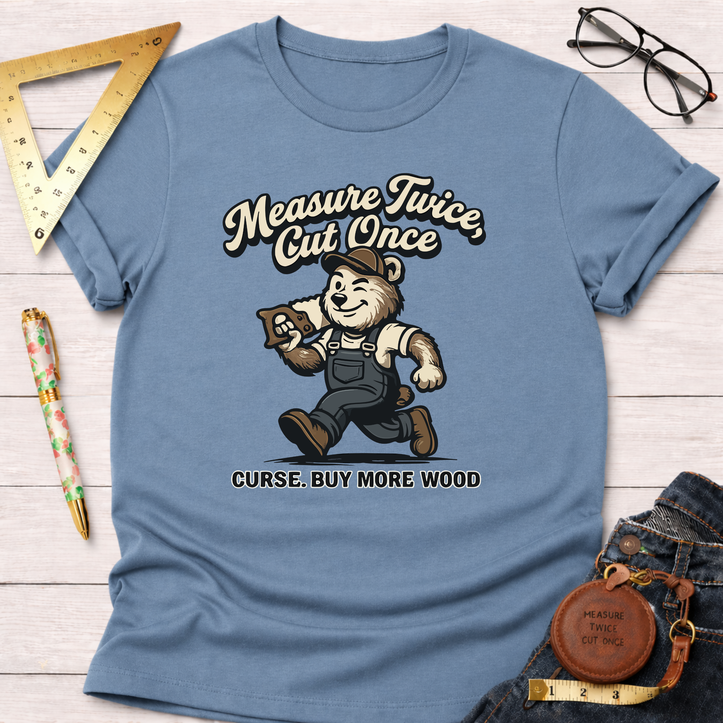 Women's funny woodworking graphic tee shirt for female woodworkers, makers, and DIY builders — Sawdust & Ponytails. Light blue cotton tee with a cartoon bear carrying wood and a saw, text "Measure Twice, Cut Once. Curse. Buy More Wood.