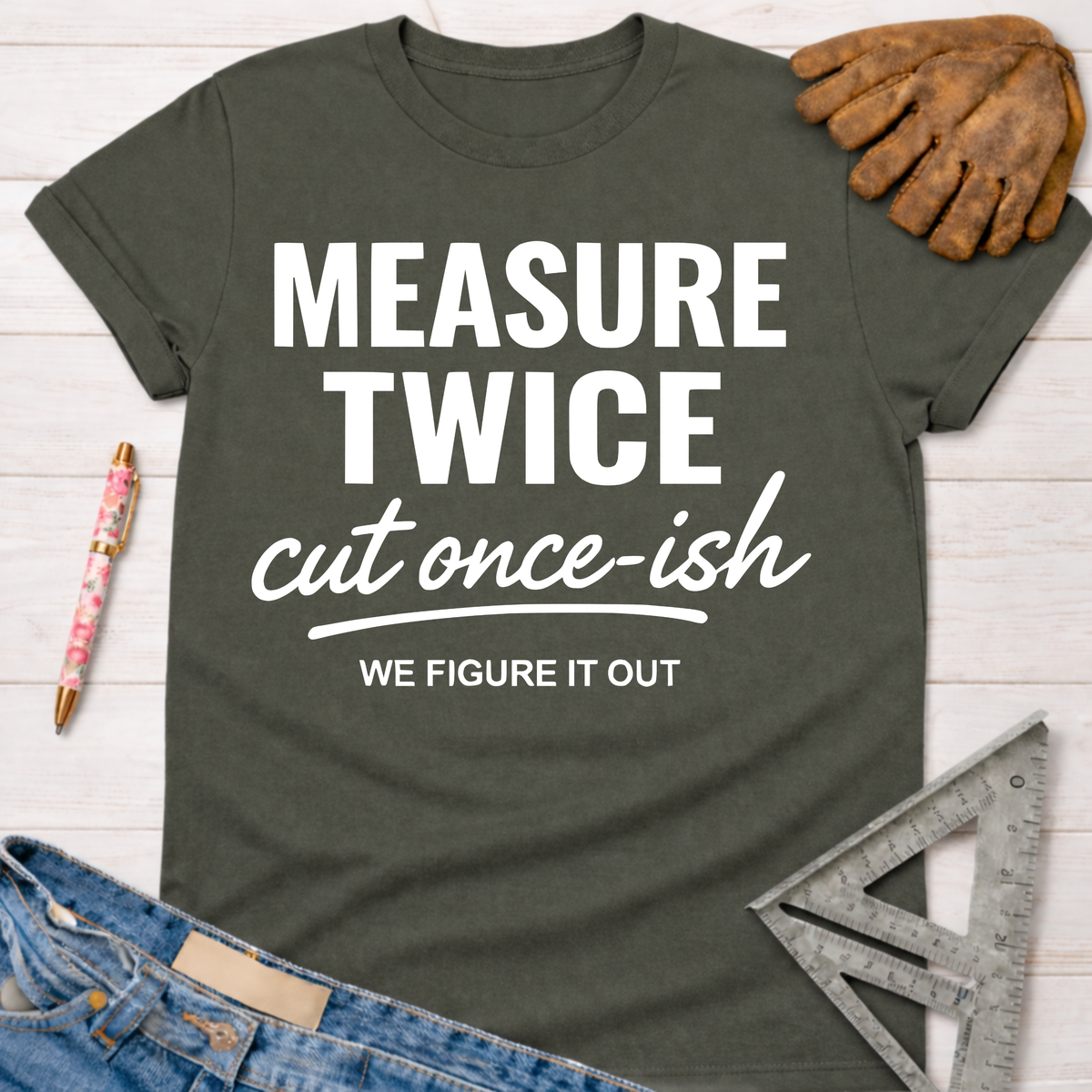 Women's funny woodworking graphic tee shirt for female woodworkers, makers, and DIY builders — Sawdust & Ponytails. Dark olive green crewneck t-shirt with "MEASURE TWICE cut once-ish WE FIGURE IT OUT" in white text.