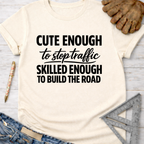 Women's funny woodworking graphic tee shirt for female woodworkers, makers, and DIY builders — Sawdust & Ponytails. Cream t-shirt with "Cute Enough To Stop Traffic Skilled Enough To Build The Road" in black text.