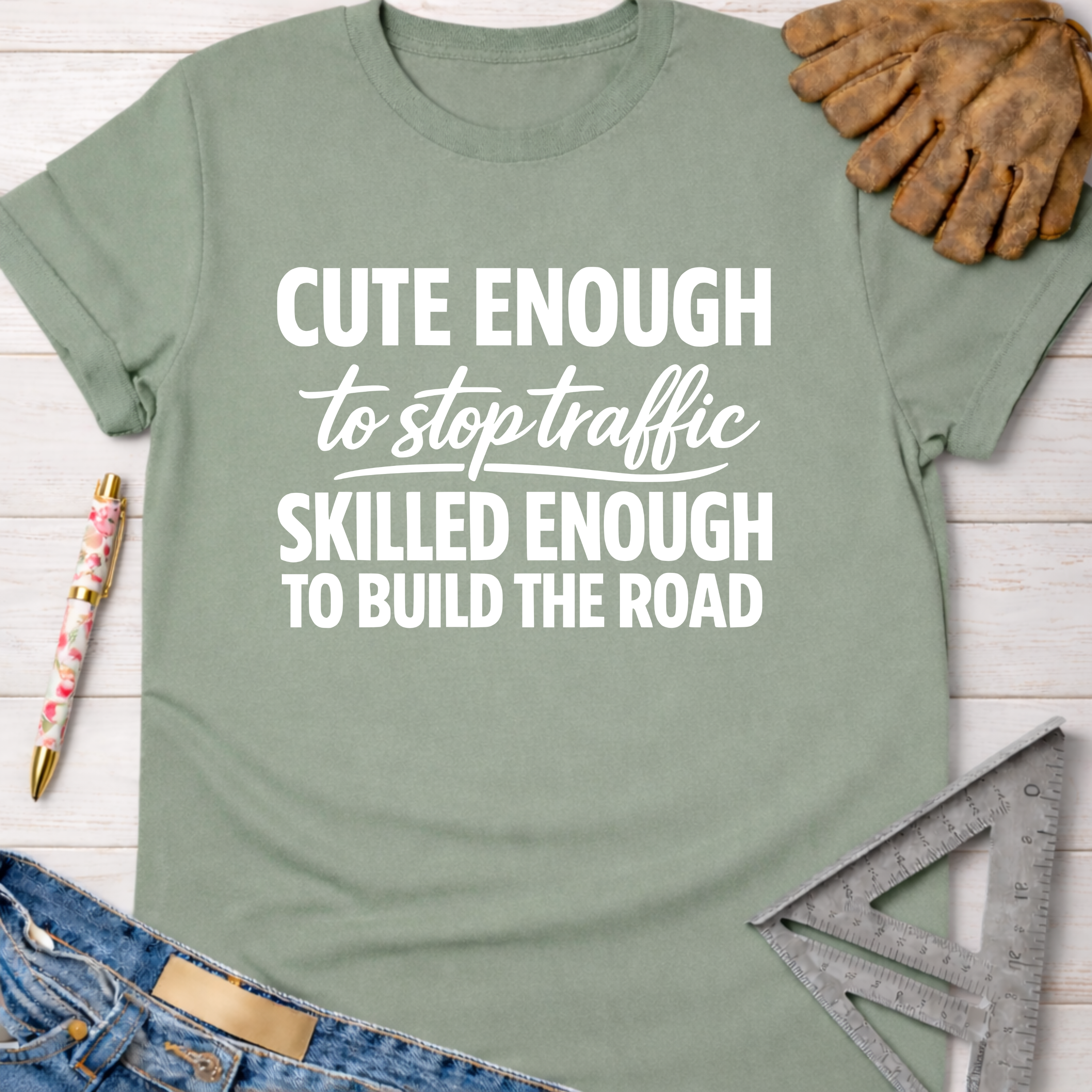 Women's funny woodworking graphic tee shirt for female woodworkers, makers, and DIY builders — Sawdust & Ponytails. Sage green cotton tee with "Cute Enough To Stop Traffic Skilled Enough To Build The Road" in white text.