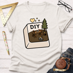 Women's funny woodworking graphic tee shirt for female woodworkers, makers, and DIY builders — Sawdust & Ponytails. Cream cotton tee with a "DIY" button graphic featuring a workbench, saw, and pine tree.