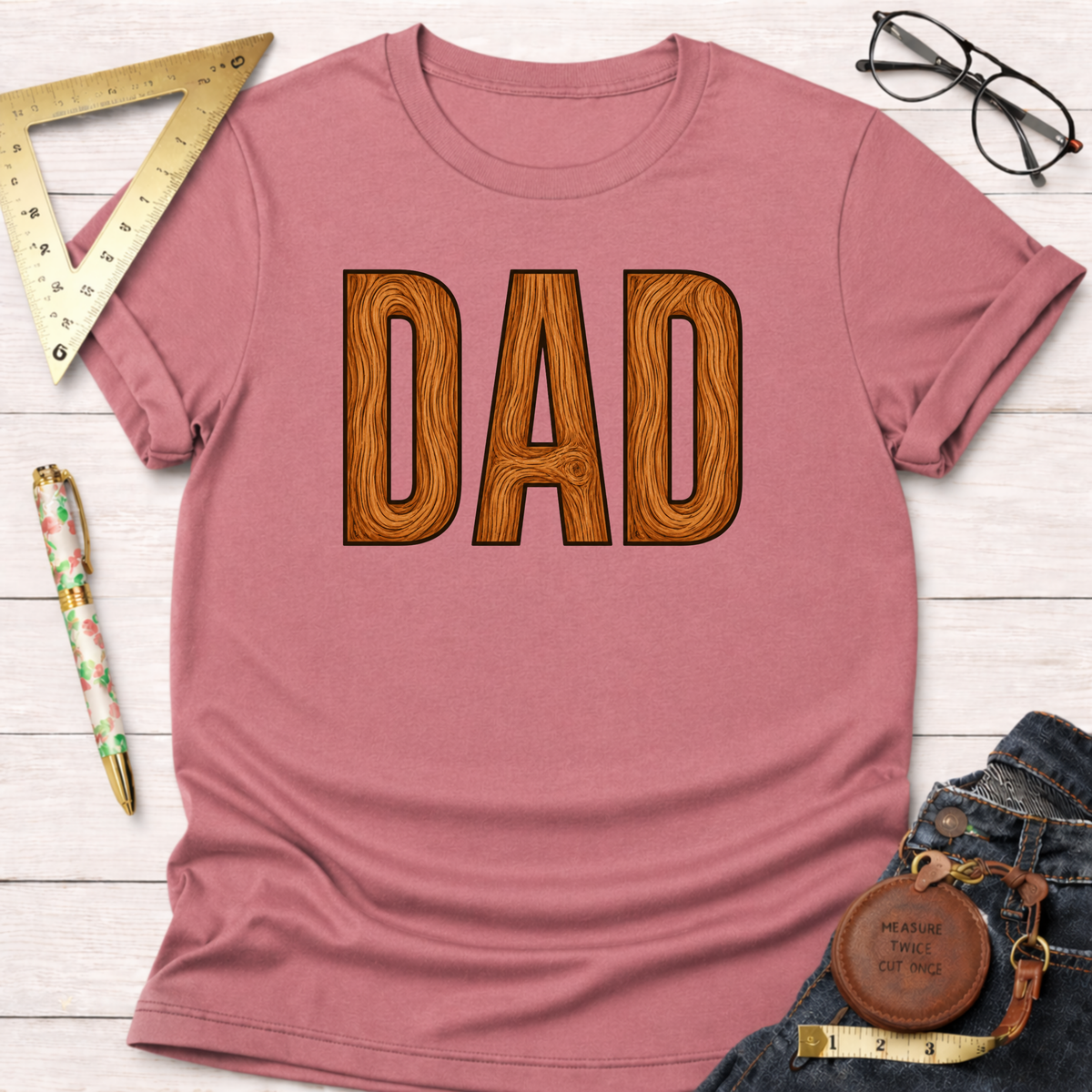 Pink short-sleeve t-shirt with "DAD" in a wood grain font, styled with jeans and woodworking tools.
