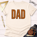Cream t-shirt with "DAD" in brown wooden letters, styled with jeans, ruler, pen, and glasses.