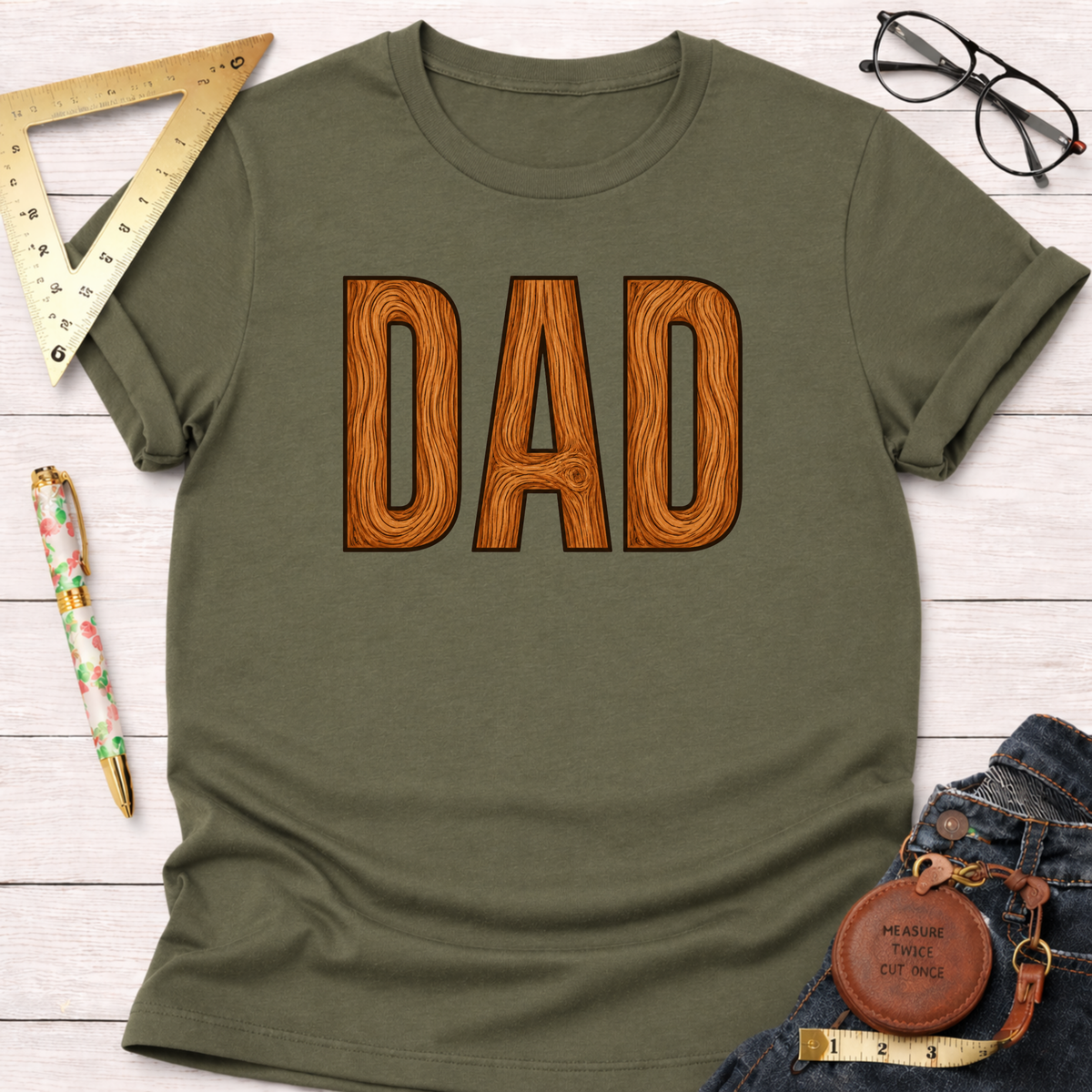 Olive green t-shirt with "DAD" in brown wooden letters, styled with jeans, ruler, and glasses.