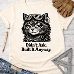 Women's funny woodworking graphic tee shirt for female woodworkers, makers, and DIY builders — Sawdust & Ponytails. Cream t-shirt with a black graphic of a cat wearing goggles and a flower, text "Didn't Ask. Built It Anyway.