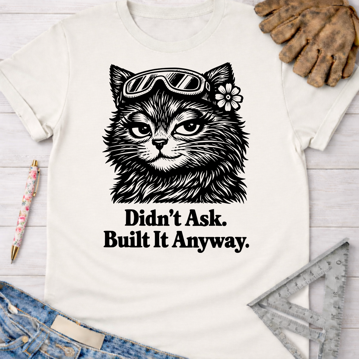 Women's funny woodworking graphic tee shirt for female woodworkers, makers, and DIY builders — Sawdust & Ponytails. White cotton tee with a black graphic of a cat wearing safety goggles and a flower, with text "Didn't Ask. Built It Anyway.