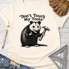 Women's funny woodworking graphic tee shirt for female woodworkers, makers, and DIY builders — Sawdust & Ponytails. Cream t-shirt with a black graphic of an opossum holding a hammer and the text "Don't Touch My Tools".