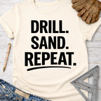 Women's funny woodworking graphic tee shirt for female woodworkers, makers, and DIY builders — Sawdust & Ponytails. Cream t-shirt with "DRILL. SAND. REPEAT." in black text.