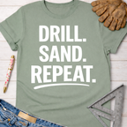 Women's funny woodworking graphic tee shirt for female woodworkers, makers, and DIY builders — Sawdust & Ponytails. Sage green cotton t-shirt with "DRILL. SAND. REPEAT." in white text.