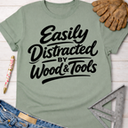 Women's funny woodworking graphic tee shirt for female woodworkers, makers, and DIY builders — Sawdust & Ponytails. Sage green cotton t-shirt with "Easily Distracted by Wood & Tools" in black script.