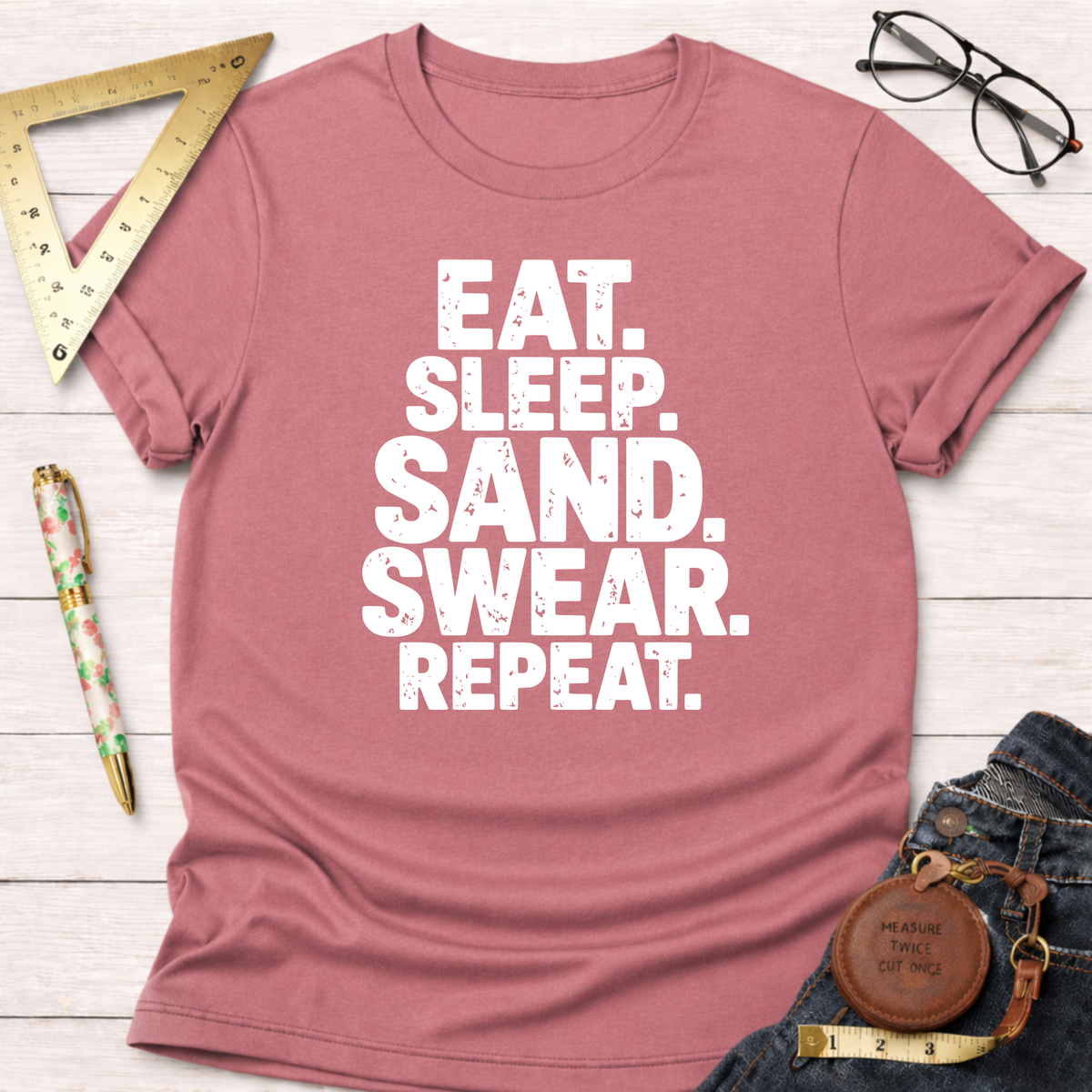 Dusty rose t-shirt with "Eat. Sleep. Sand. Swear. Repeat." in white text, styled with jeans and tools.