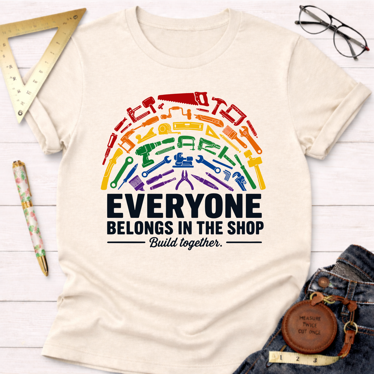 Cream unisex t-shirt with a rainbow arc of diverse tools and "Everyone Belongs In The Shop Build together." text.