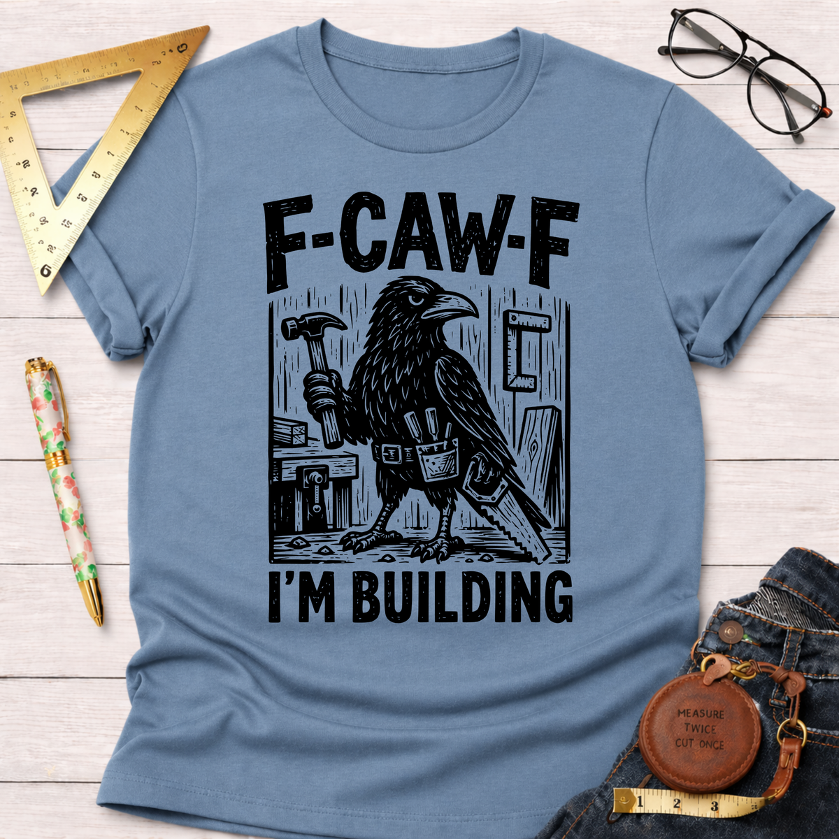 Women's funny woodworking graphic tee shirt for female woodworkers, makers, and DIY builders — Sawdust & Ponytails. Light blue cotton tee with a black crow holding a hammer and "F-CAW-F I'M BUILDING" text.