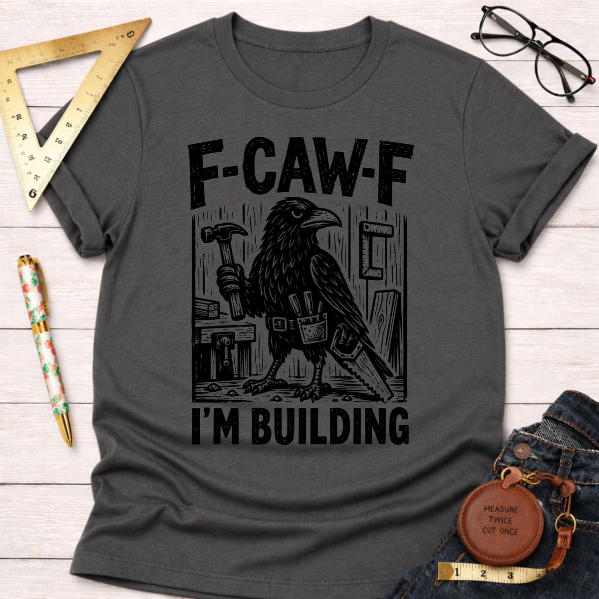 Women's funny woodworking graphic tee shirt for female woodworkers, makers, and DIY builders — Sawdust & Ponytails. Dark gray cotton tee with a black crow holding a hammer and saw, "F-CAW-F I'M BUILDING" text.