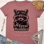 Women's funny woodworking graphic tee shirt for female woodworkers, makers, and DIY builders — Sawdust & Ponytails. Mauve cotton tee with a black raccoon in sunglasses holding a tool and "Feral Builder" text.