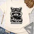 Women's funny woodworking graphic tee shirt for female woodworkers, makers, and DIY builders — Sawdust & Ponytails. Cream t-shirt with a black graphic of a raccoon in sunglasses holding a hammer, text "Feral Builder".