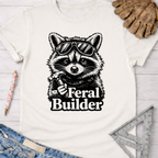 Women's funny woodworking graphic tee shirt for female woodworkers, makers, and DIY builders — Sawdust & Ponytails. Cream t-shirt with a cool raccoon in sunglasses holding a hammer and "Feral Builder" text.