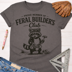 Women's funny woodworking graphic tee shirt for female woodworkers, makers, and DIY builders — Sawdust & Ponytails. Dark gray cotton tee with "Feral Builder's Club" text and a raccoon wearing goggles, holding a hammer.