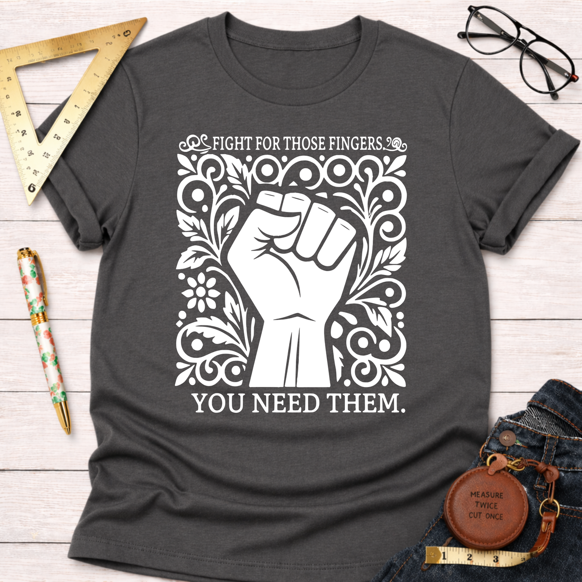 Charcoal gray cotton t-shirt with a white graphic of a raised fist surrounded by ornate floral designs and text: "FIGHT FOR THOSE FINGERS. YOU NEED THEM.